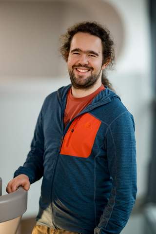 Porträt eines lächelnden Mannes mit dunklen, lockigen Haaren und Bart, der eine blaue Jacke mit rotem Brusttaschen-Akzent trägt.