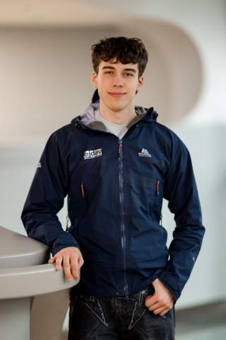 Porträt eines jungen Mannes mit kurz gewellten dunklen Haaren, der eine blaue Outdoor-Jacke trägt und entspannt an einem hellen Geländer lehnt.