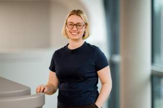 Junge Frau mit blonden Haaren und Brille lächelt stehend in einem modernen Innenraum, eine Hand lehnt an einem grauen Geländer.