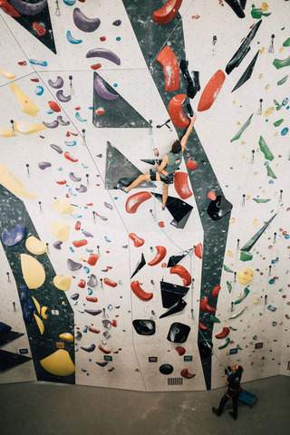Person klettert an einer bunt gestalteten Indoor-Kletterwand mit verschiedenen Griffen, während unten eine weitere Person zuschaut.
