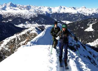 Zwei Skifahrer mit Ausrüstung wandern auf einem schmalen, schneebedeckten Berggrat mit schneebedeckten Bergen im Hintergrund.