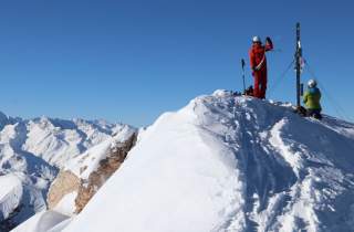 Zwei Skifahrer mit Helmen stehen und knien auf einem schneebedeckten Berggipfel, im Hintergrund sind verschneite Bergketten unter klarem blauem Himmel zu sehen.