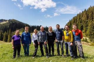 Acht Menschen stehen lächelnd nebeneinander auf einer Wiese in den Bergen, umgeben von Nadelwäldern und blauem Himmel.