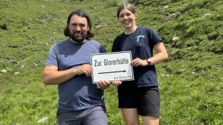 Zwei Menschen beim wandern halten Schild "Zur Glorerhütte" hoch