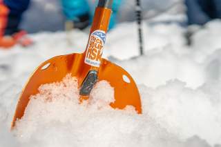 Eine orangene Schaufel mit Check-Your-Risk Sticker steckt im Schnee