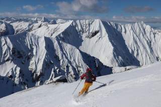 Ein Skifahrer in roter Jacke und gelber Hose fährt einen verschneiten Hang hinunter, umgeben von einer Berglandschaft mit schroffen, schneebedeckten Gipfeln. Der klare Himmel und die Berge im Hintergrund vervollständigen die Szenerie.