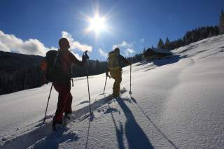 Zwei Skibergsteiger besteigen einen schneebedeckten Hang. Im Hintergrund ist eine Berghütte zu sehen