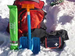 Rotes Rucksack-Set mit Lawinensonde in grünem Etui, blauer Schaufel mit orangefarbenem Griff und Erste-Hilfe-Set im Schnee.