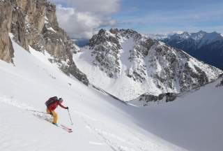 Ein Skifahrer in roter Jacke und gelber Hose fährt einen steilen, verschneiten Hang hinunter, flankiert von imposanten Felswänden. Im Hintergrund erstreckt sich eine spektakuläre Berglandschaft mit schneebedeckten Gipfeln unter einem leicht bewölkten Himmel.