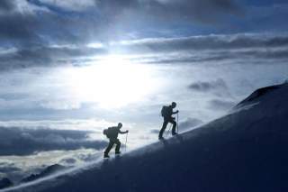 Zwei Personen mit Rucksäcken und Trekkingstöcken wandern einen schneebedeckten Berghang hinauf, während der Himmel bewölkt ist und die Sonne durchscheint.