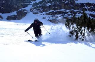Ein Skifahrer fährt eine verschneite Piste hinunter, während er eine scharfe Kurve nimmt und Schnee aufwirbelt. Im Hintergrund sind schneebedeckte Felsen und einige Bäume zu sehen.