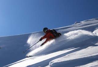 Ein Skifahrer in roter Jacke und schwarzem Helm fährt unter blauem Himmel einen verschneiten Hang hinunter.