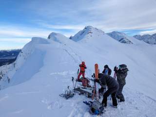 Eine Gruppe von Skifahrern und Snowboardern bereitet sich auf einer schneebedeckten Bergkuppe unter einem leicht bewölkten Himmel auf die Abfahrt vor.