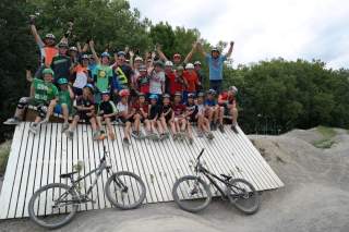 Eine große Gruppe Kinder und zwei Erwachsene mit Fahrradhelmen sitzen und stehen auf einer schrägen Holzrampe, vor ihnen stehen zwei Fahrräder, im Hintergrund sind Bäume zu sehen.