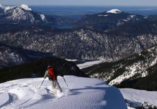 Ein Skifahrer in grauer Kleidung und mit rotem Rucksack fährt einen schneebedeckten Hang hinunter, umgeben von bergigem Gelände und unter einem klaren Himmel.