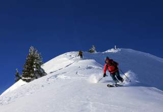 Zwei Skifahrer fahren bei strahlend blauem Himmel eine schneebedeckte Piste hinunter. Der vordere Skifahrer trägt eine rote Jacke und blaue Mütze, während der hintere Skifahrer eine gelbe Jacke trägt. Einige verschneite Bäume sind im Hintergrund zu sehen.
