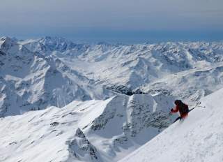 Ein Skifahrer fährt einen steilen hang hinab. Im Hintergrund ist eine beeindruckende, mit Schnee bedeckte Bergkulisse zu sehen