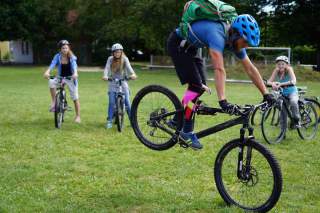 Ein Mann führt auf einer Wiese einen Radtrick mit einem Mountainbike vor, während drei Mädchen mit Fahrrädern zuschauen. Alle tragen Fahrradhelme.