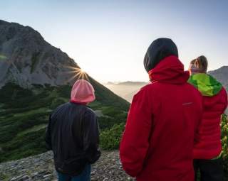 Drei Personen in Outdoor-Bekleidung stehen auf einem Berg und blicken in Richtung Sonnenaufgang hinter einem anderen Berg.