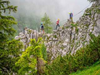 Vier Kletterer mit Helmen und Sicherungsausrüstung überqueren eine felsige Bergkante, umgeben von grünen Bäumen und Nebel.