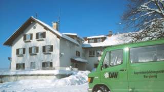 Schneebedecktes Gebäude hinter einem grünen Fahrzeug mit der Aufschrift „DAV Jugendbildungsstätte Burgberg“ an einem sonnigen Wintertag.