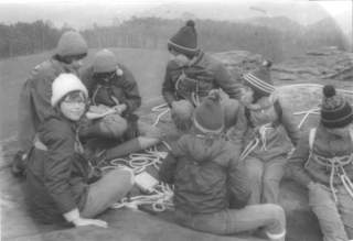 Eine Gruppe Kinder mit Winterkleidung und Strickmützen sitzt auf einem Felsen im Freien, umgeben von Seilen, und scheint für eine Kletteraktivität vorbereitet zu sein.
