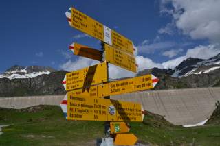 Wegweiser mit gelben Schildern und Wanderzielen sowie Gehzeiten vor einer Staumauer und schneebedeckten Bergen unter blauem Himmel.