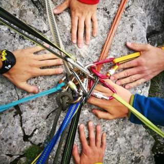 Ein Bild von mehreren Händen, die sich auf einem Felsen treffen und verschiedene Kletterseile und Karabiner sichern