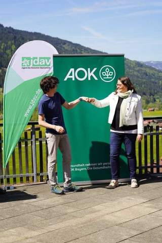 Ein Mann und eine Frau geben sich die Hand vor Bannern der JDAV und AOK, im Hintergrund sind Berge zu sehen.