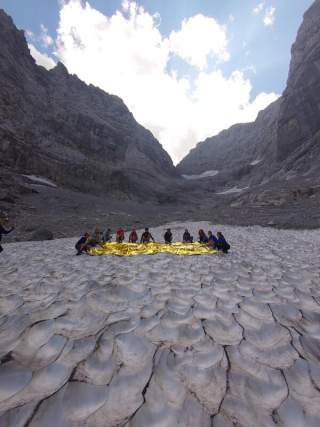 Eine Gruppe von Menschen sitzt auf einem Gletscher, umgeben von hohen Bergen, und hält eine goldene Rettungsdecke ausgebreitet.