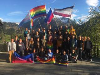 Gruppenbild des Queerfeldein 2019 auf einer Terasse im Hintergrund die Berge und verschiedene Flaggen der Queeren Community