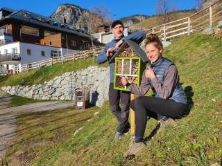 Zwei Personen zeigen draußen bei Sonnenschein einen kleinen, grünen Insektenhotel-Kasten auf einem Holzpfahl, im Hintergrund sind Häuser und ein Berg zu sehen.