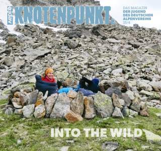 Zwei Menschen lehnen entspannt in einer Sitzmulde aus Steinen vor einem steinigen Berghang, umgeben von Gras. Im Hintergrund ist der Schriftzug „KNOTENPUNKT.“ und „INTO THE WILD“ zu sehen.