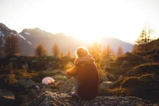 Wanderin sitzt auf Fels und umschlingt Beine mit den Armen, guckt in die Abendsonne